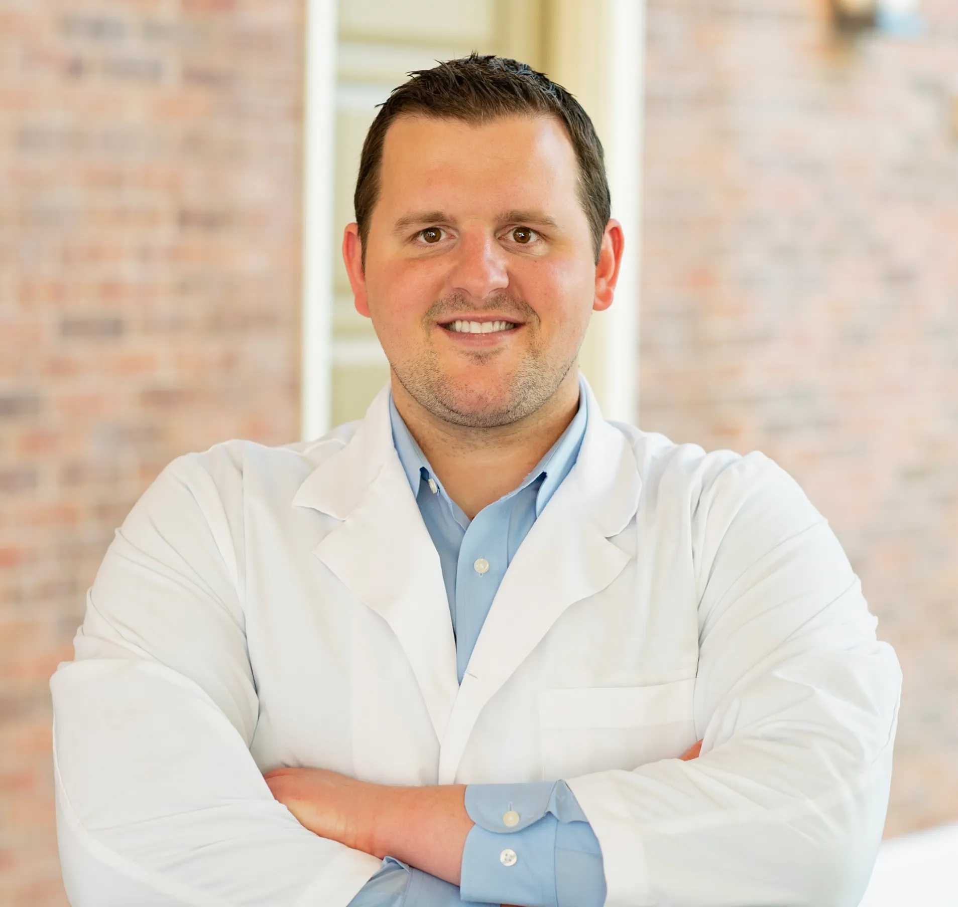 Smiling man in white lab coat with blue shirt standing with arms crossed in front of a brick wall.